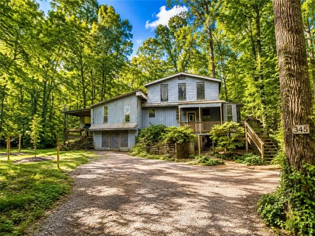 Wooded property with a gravel driveway, mature trees, and a multi-level wood-sided residence featuring an elevated porch and exterior stairs