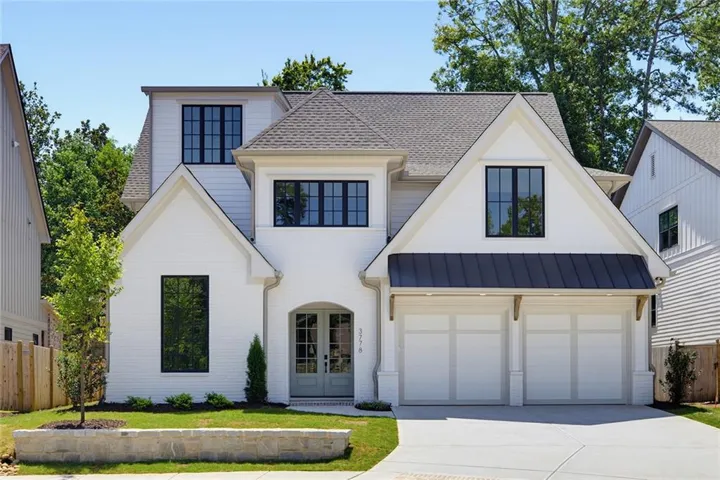 White brick exterior with black framed windows, two-car garage with overhead metal awning, arched double-door entry, and a stone retaining wall