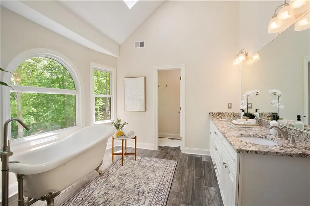 The spa-like primary bathroom offers a serene escape, featuring a freestanding soaking tub perfectly positioned beneath sunlit windows with peaceful wooded views.