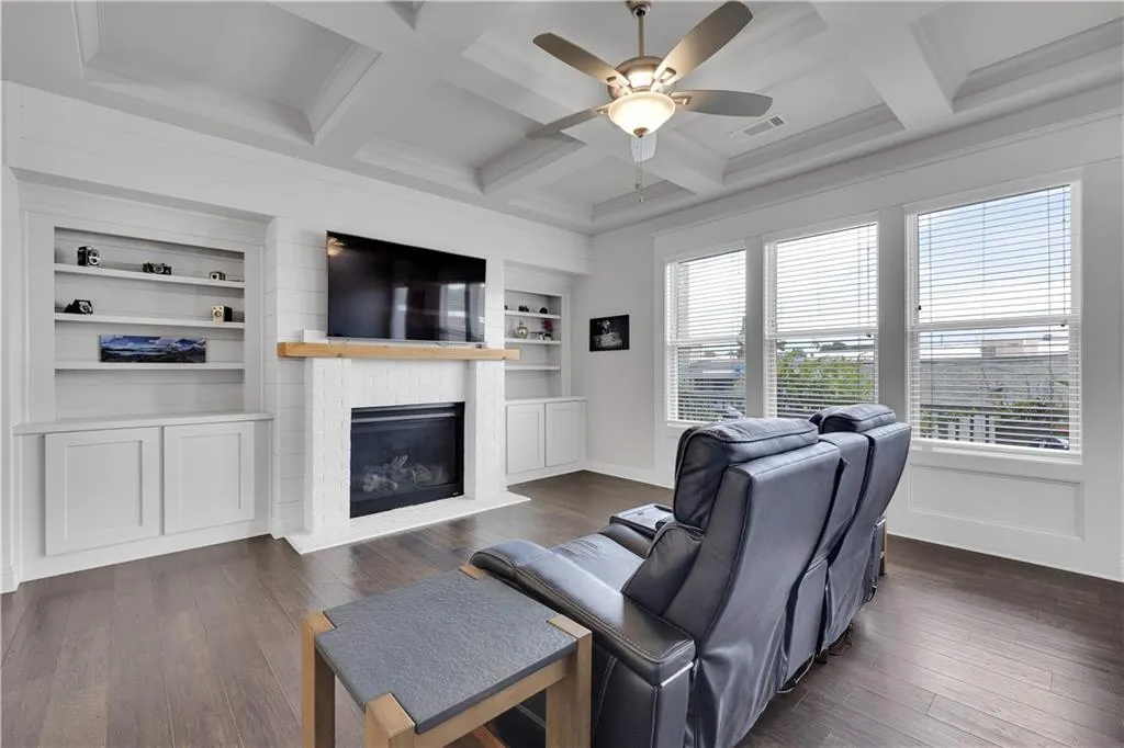 Living room with a brick fireplace, dark wood-style flooring, beam ceiling, ceiling fan, and built in shelves