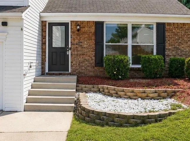 Brick facade, concrete steps, dark entry door with glass panel, white siding, and layered landscaping with retaining walls