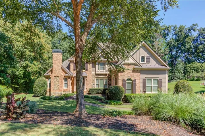 Stone and brick facade with shake siding accents, shuttered windows, and a prominent brick chimney, set on a landscaped lot with mature trees