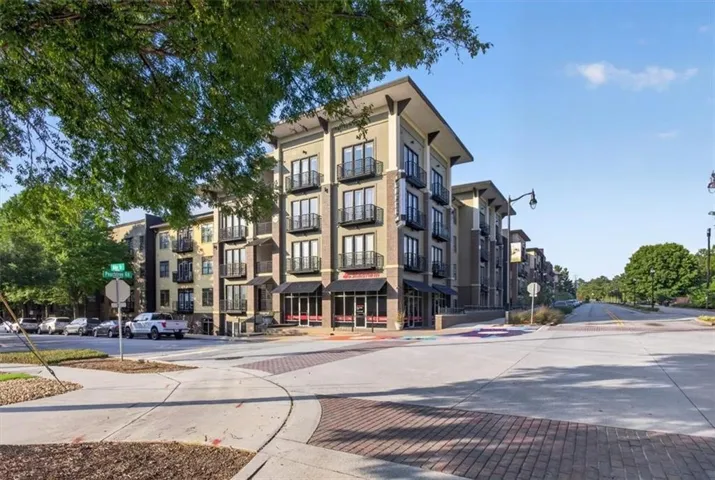 Multi-story residential building featuring balconies with black metal railings, commercial storefronts on the ground level, and a paved walkway with brick accents