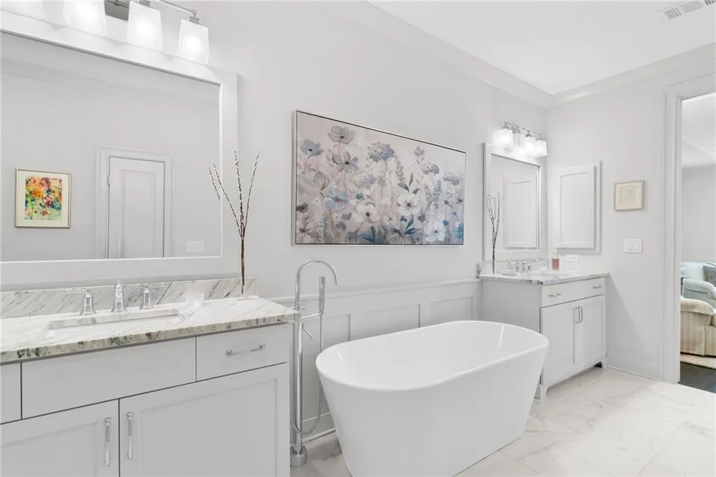Freestanding white soaking tub with a floor-mounted chrome faucet, dual white cabinetry vanities with light-toned stone countertops, integrated undermount sinks, and wall-to-wall framed mirrors