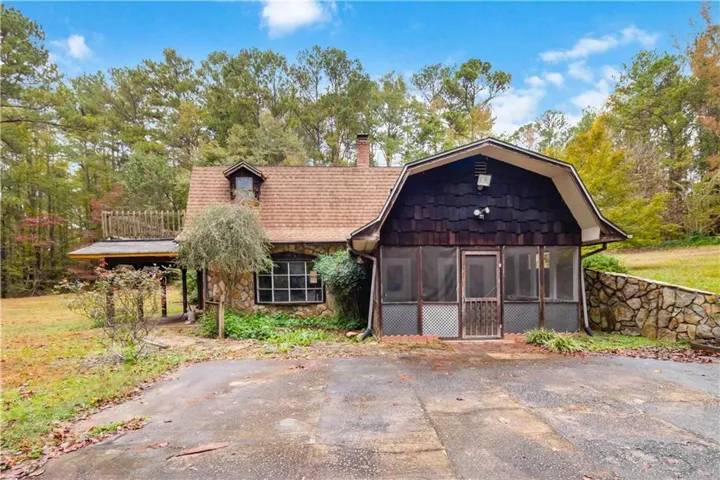 View of front facade featuring a gambrel roof, a chimney, stone siding, a sunroom, and roof with shingles