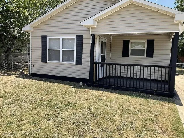 Exterior featuring light-toned horizontal siding, dark window shutters, a covered porch with dark railings and flooring, and a fenced side yard