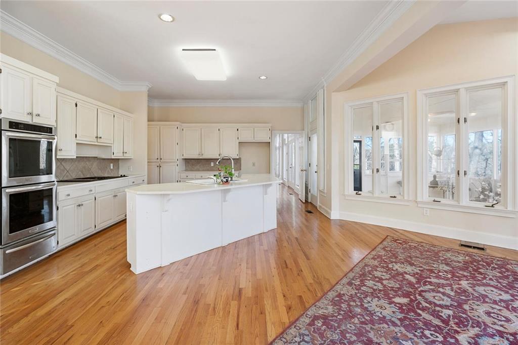 Kitchen featuring stainless steel double oven, white cabinets, light wood-type flooring, backsplash, and a center island with sink