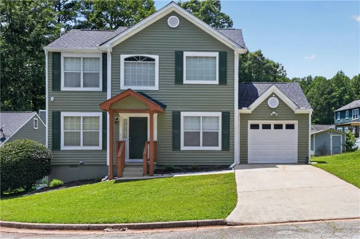 Two-story residence featuring green siding, a covered entry porch, a single-car garage, multiple windows with dark shutters, and a sloped, grassed front yard