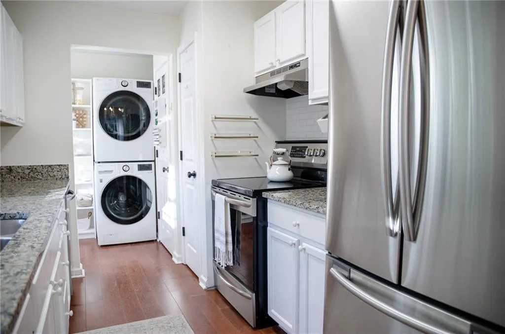 Kitchen with appliances with stainless steel finishes, white cabinetry, light stone countertops, under cabinet range hood, and stacked washing machine and dryer