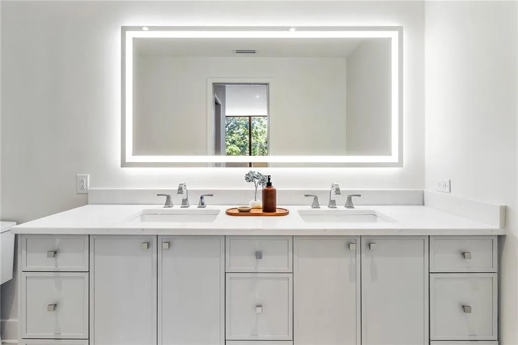 Dual vanity featuring a white solid-surface countertop, integrated rectangular sinks, polished chrome widespread faucets, and light gray shaker-style cabinetry with square hardware, illuminated by a large rectangular LED-backlit mirror