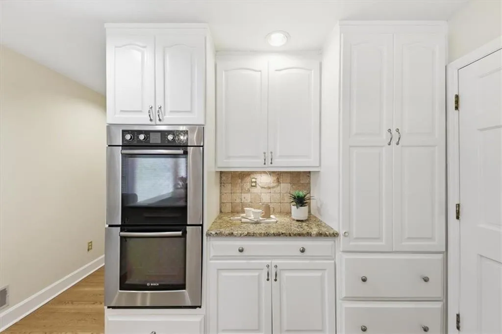 Kitchen with stainless steel double oven, white cabinetry, light stone countertops, decorative backsplash, and light wood-style flooring