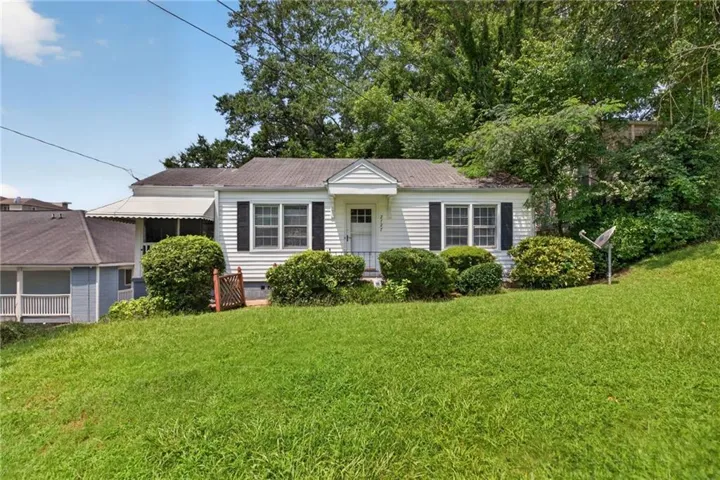 White siding residence with dark window shutters, a covered porch entrance, an awning-covered side porch, a sloping green lawn, and mature tree landscaping