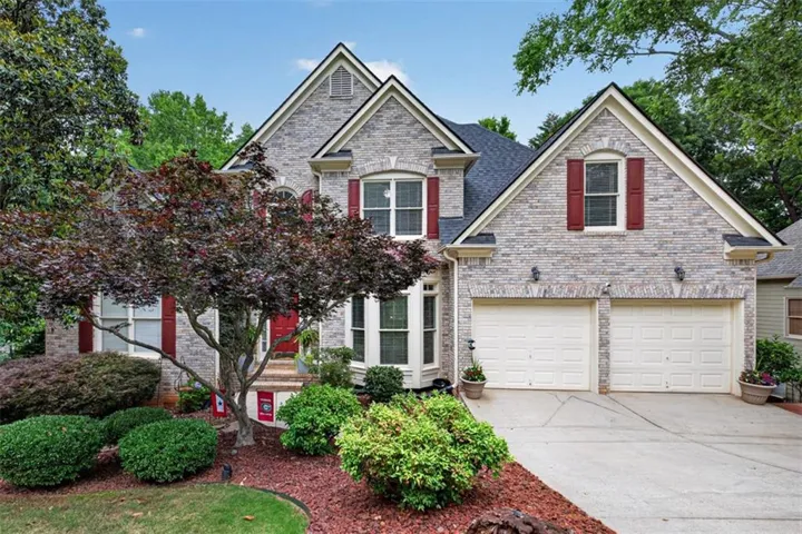Brick exterior with a dark shingle roof, two-car garage, red window shutters, and mature landscaping