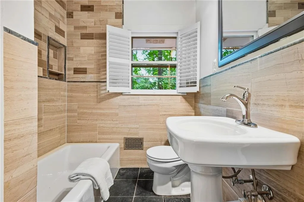 Full-height neutral stone wall tiling, white pedestal sink with polished chrome faucet, built-in tiled shower niche, operable interior shutters, and dark stone floor tiling