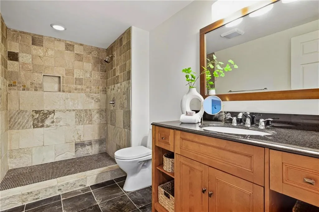 Bathroom featuring a walk-in shower with stone tile surround and built-in niche, dark stone slab countertop, wood cabinetry with open shelving, and a framed mirror