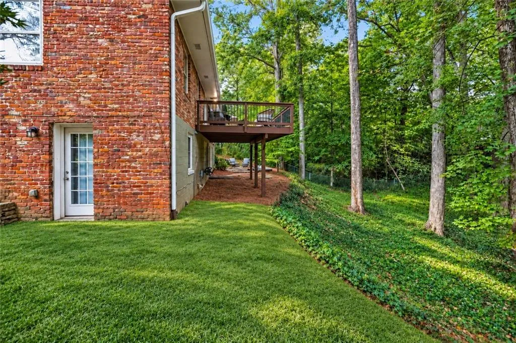 Multi-level brick exterior featuring an elevated wood-railing deck, manicured lawn, sloped landscape, and mature trees