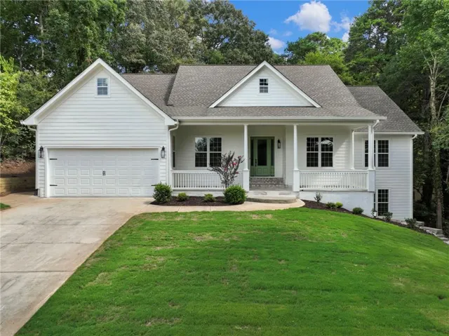 White siding exterior with a prominent covered front porch, an attached garage, a verdant lawn, and mature trees in the background