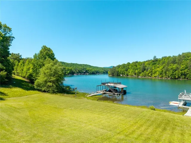 Expansive waterfront property featuring a manicured lawn, direct lake access, a private covered boat dock, an additional uncovered dock structure, and mature surrounding tree lines