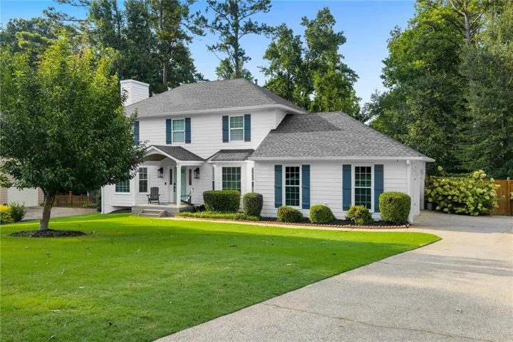 White exterior siding with blue shutters, covered front porch, asphalt shingle roof, concrete driveway, and mature landscaping