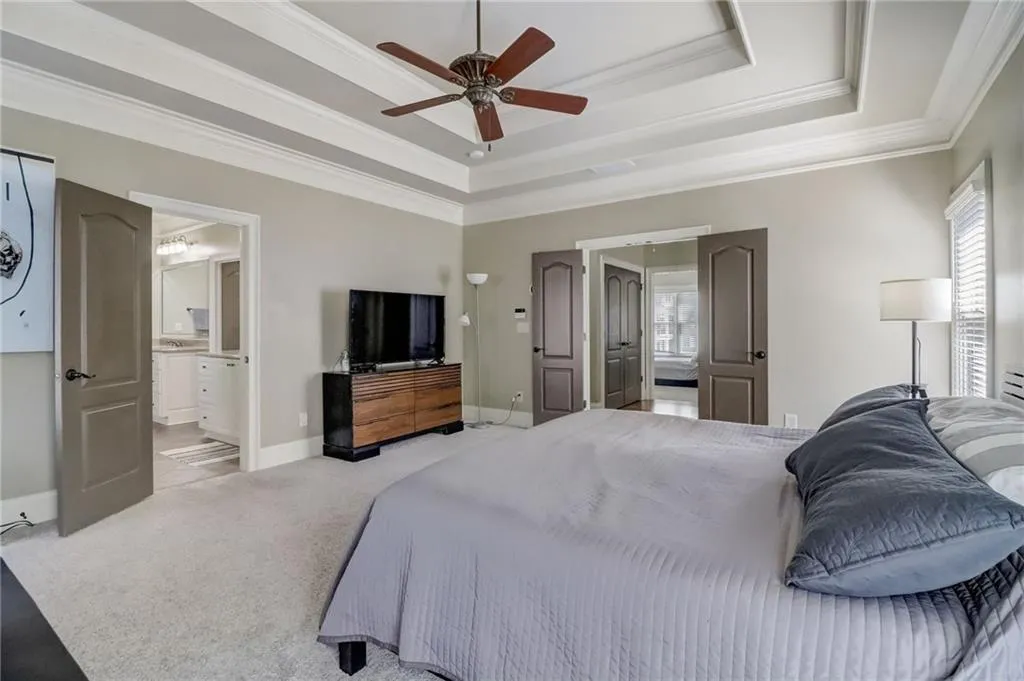 Carpeted bedroom with ornamental molding, ceiling fan, ensuite bath, and a raised ceiling