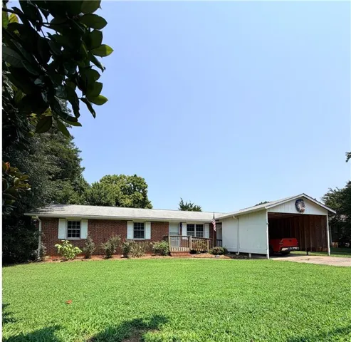 Brick exterior residence featuring a front lawn, white window shutters, a wooden front porch with a ramp, and an attached white carport