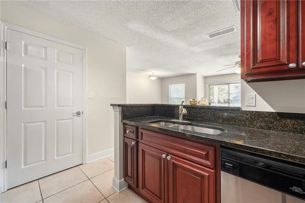 Kitchen with stainless steel dishwasher, dark brown cabinets, light tile patterned floors, dark stone countertops, and a textured ceiling