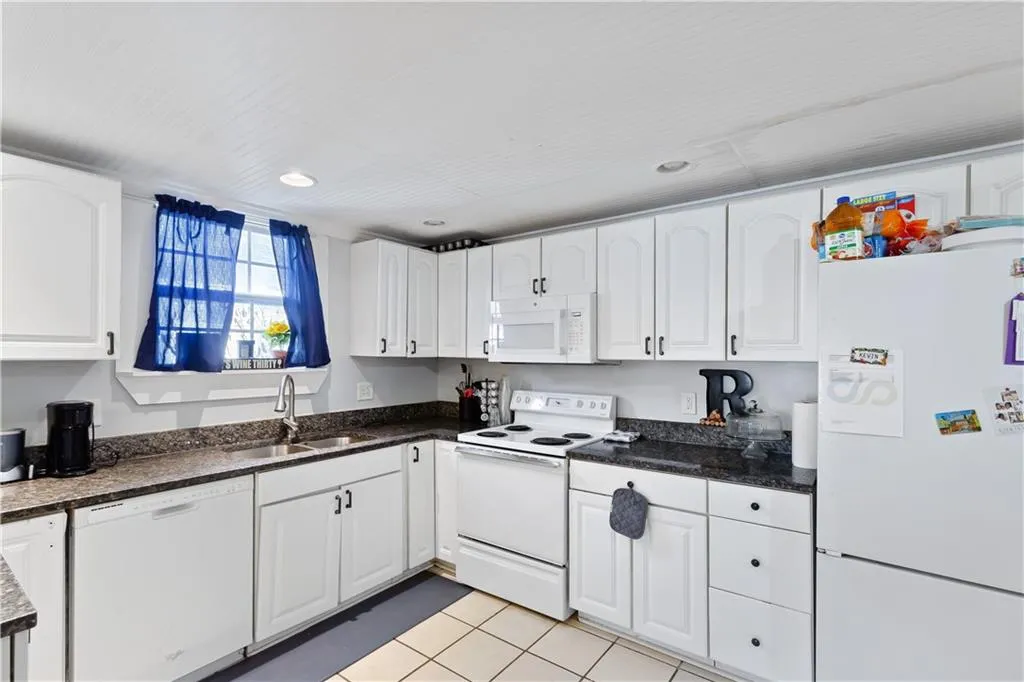Kitchen featuring light tile flooring, dark stone countertops, white appliances, sink, and white cabinetry