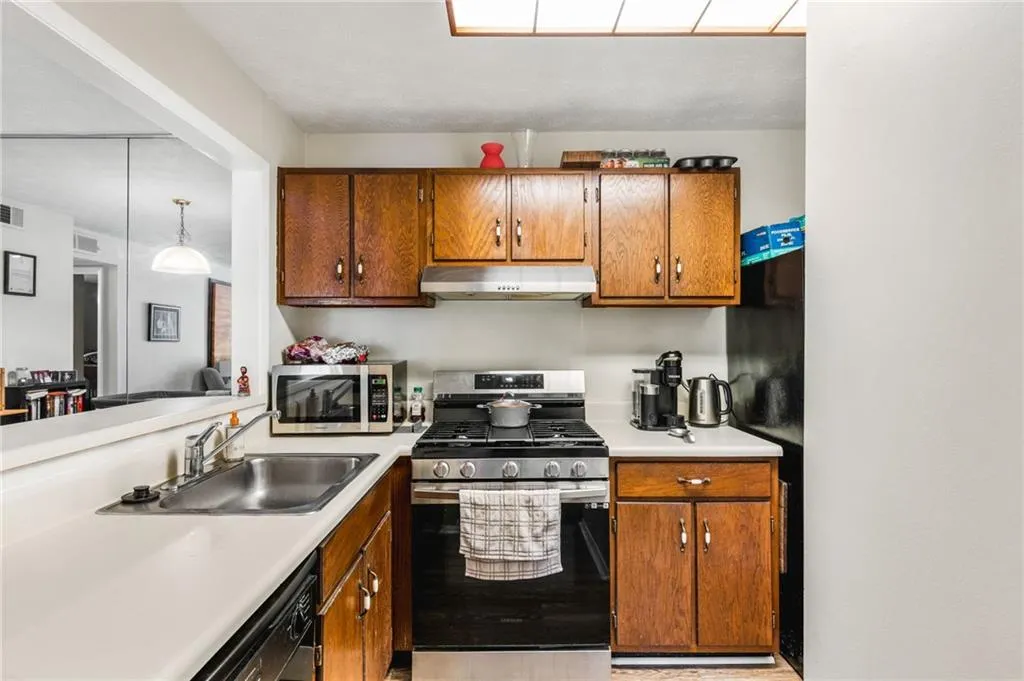 Kitchen featuring under cabinet range hood, light countertops, brown cabinetry, black appliances, and a sink