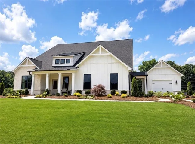 Contemporary farmhouse exterior featuring white board and batten siding, light brick accents, a dark shingle roof, a covered front porch with white columns, and a two-car garage with a white overhead door