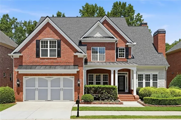 Brick facade residence featuring a covered front entry, arched garage doors, a shingle roof, established landscaping, and a paved driveway