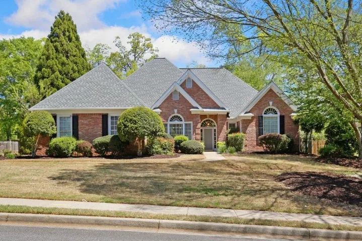 Brick exterior featuring a gray shingle roof, arched transom windows, black louvered shutters, and established perimeter landscaping