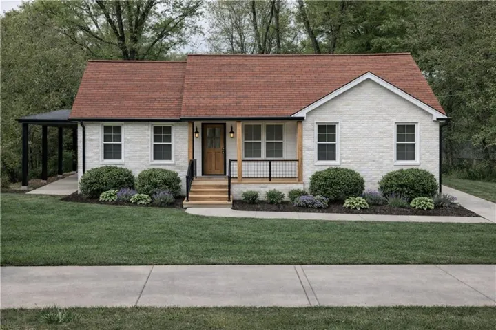 Light-colored brick exterior with a contrasting red roof, a covered entry porch, a wood-finish front door, a covered carport, and a manicured lawn with mature landscaping