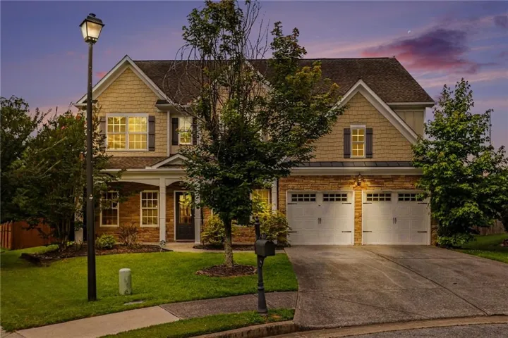 Two-story residence featuring a two-car garage with carriage-style doors, stone and shingle siding, a covered entry porch with columns, and a well-maintained lawn with mature trees