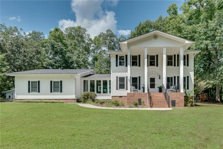 House front featuring white siding exterior with black shutters, mature tree surroundings, full-length front porch columns, front brick steps with black railings, and an extra sunroom extension