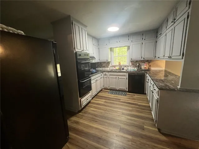 U-shaped kitchen featuring wood-finish flooring, light-colored cabinetry with dark hardware, dark countertops, built-in double oven, and a mosaic tile backsplash