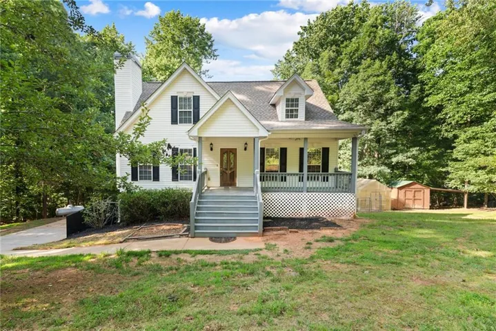 Traditional-style home featuring white siding, dark shutters, a covered front porch with steps, a detached storage shed, and mature trees surrounding the property