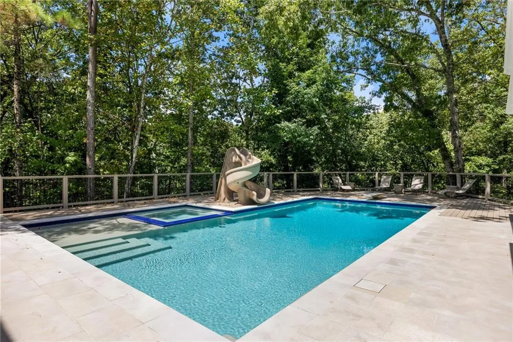 Outdoor pool featuring an integrated spa, a built-in water slide, surrounding paved decking, and a perimeter fence