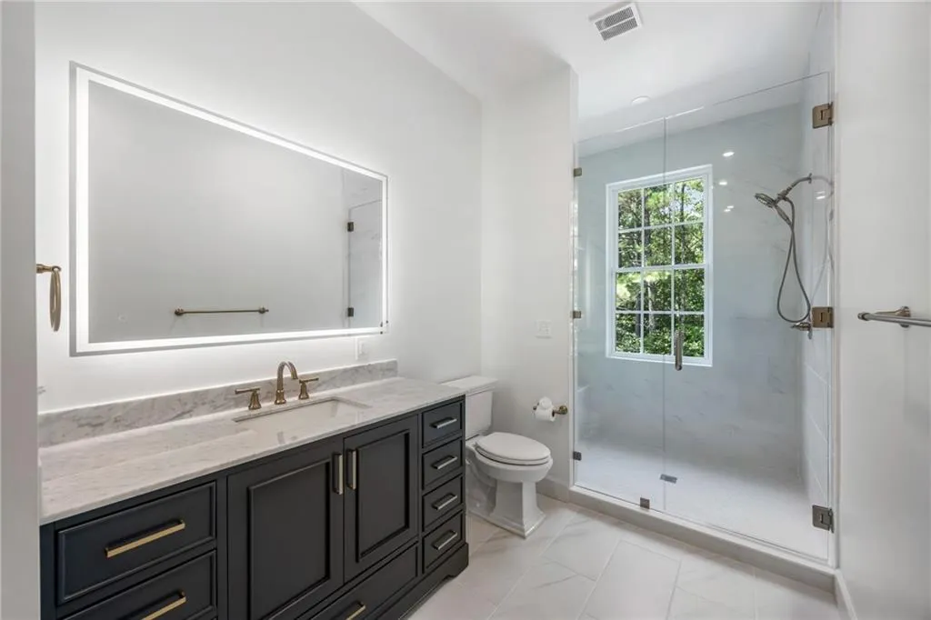 Illuminated vanity mirror, dual-tone vanity with ample storage, integrated single-basin sink, glass-enclosed shower, and a large shower window