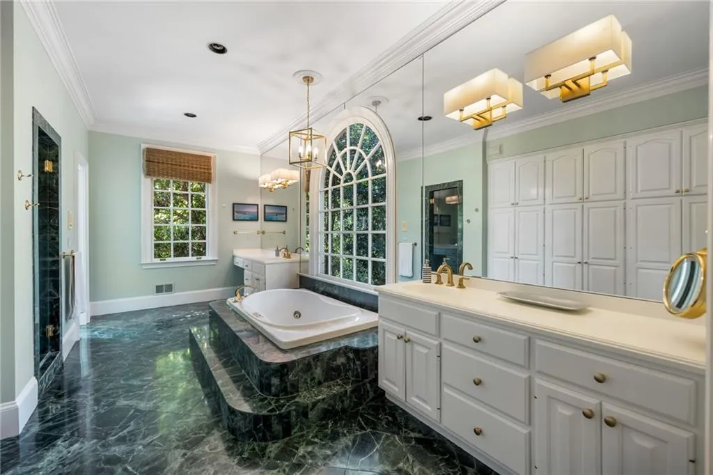Recessed soaking tub with marble surround, an arched window, a built-in vanity with white cabinetry, and extensive full-wall mirrored surfaces