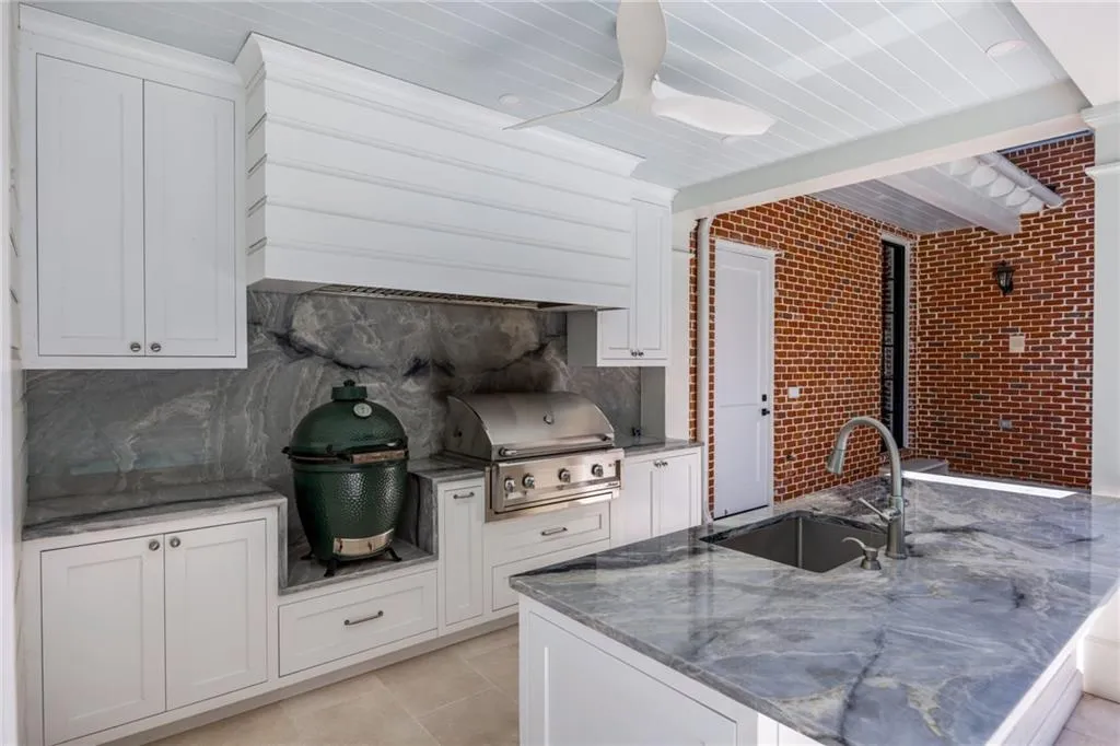 Outdoor kitchen featuring light cabinetry, integrated stainless steel grill, stone countertops, a ceiling fan, and exposed brick walls