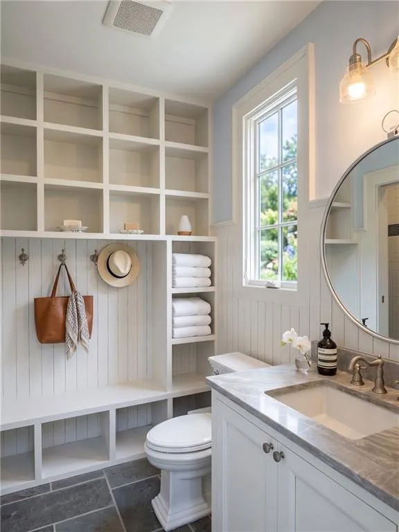 Built-in shelving, white vanity with stone countertop, undermount sink, circular mirror, and vertical wall paneling