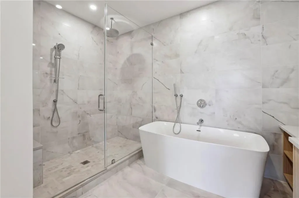 Freestanding soaking tub, frameless glass shower enclosure, rain shower head, handheld shower fixture, and large format wall tile