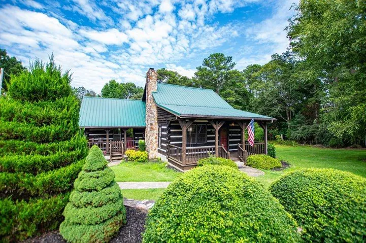 Log cabin exterior featuring a green metal roof, stone chimney, wood porch, and surrounding mature landscaping