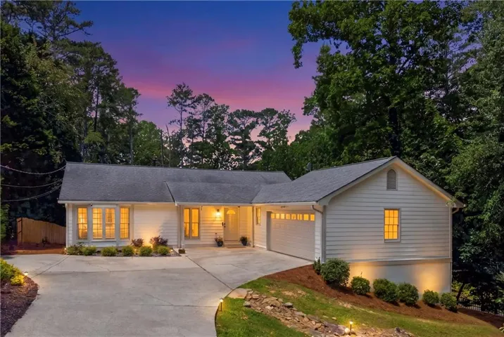 White siding residence featuring a two-car garage, mature tree backdrop, concrete driveway with integrated lighting, and a front entry with accent lighting