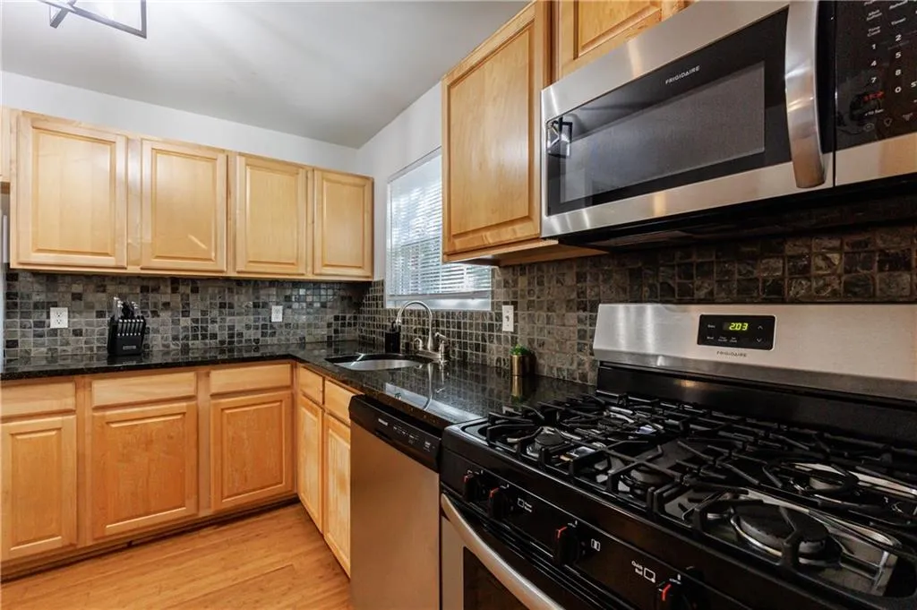 Kitchen featuring appliances with stainless steel finishes, tasteful backsplash, light wood finished floors, dark stone countertops, and light brown cabinetry