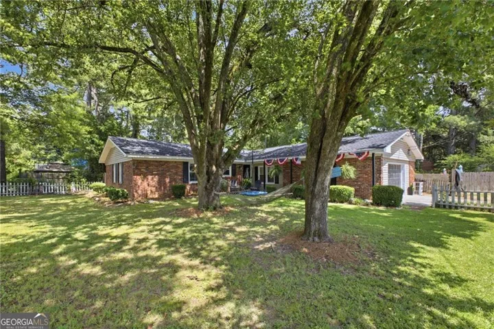 Brick exterior with light-colored siding accents, mature trees providing natural shade, a gabled roofline, and a partial picket fence