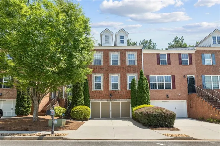 Brick townhouse facade featuring a two-car garage, multiple white-framed windows, twin dormer windows, and mature landscaping