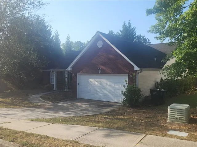 Brick home featuring a two-car garage with white paneled door, concrete driveway, and mature landscaping with evergreen trees throughout the surrounding property