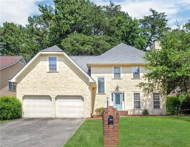 Stucco exterior with a light blue front door, two-car garage, gray shingle roof, and a mature treed backdrop