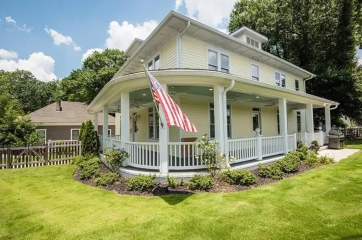 Expansive wraparound porch with white balusters, light yellow siding, multiple windows, and a manicured lawn